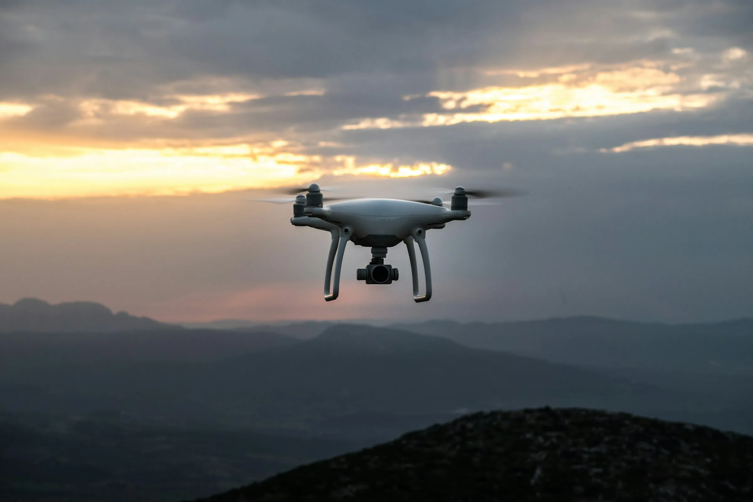 White drone in flight above a mountain valley at sunset